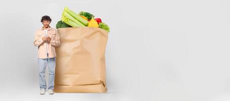 A customer stands holding a phone next to a large paper bag filled with fresh groceries. This reflects current trends in shopping and delivery during isolated times. photo