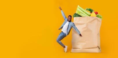 A happy customer, dressed casually, celebrates after shopping at a grocery store. The bright yellow background enhances the freshness of the vegetables in the paper bag. photo
