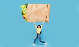 A shopper proudly holds a large paper bag filled with vibrant vegetables and fruits. The backdrop features a bright blue color, emphasizing the excitement of grocery shopping. photo