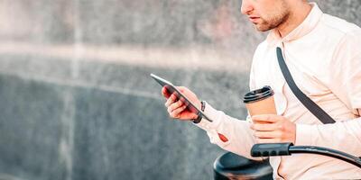 Work and study online, urgent task and eco transport. Serious handsome millennial man sits on bicycle with cup of takeaway coffee and reads message on tablet, on gray street wall background, outdoor photo
