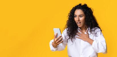 Omg. Shocked Brunette Lady Looking At Smartphone Screen With Surprised Face Expression, Emotionally Reacting To News, Reading Message, Got Unexpected Offer, Standing Over Yellow Background, Free Space photo