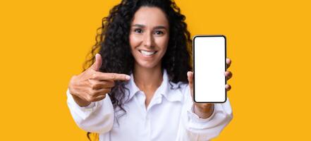 Mockup Image Of Brunette Woman Pointing At Smartphone With Blank White Screen, Demonstrating Free Place For Design Or Advertisement, Recommending App Or Website, Posing On Yellow Studio Background photo
