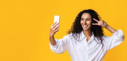 Positive Millennial Lady Taking Selfie On Smartphone And Showing Peace Gesture At Camera, Playful Brunette Woman Having Fun While Making Photo, Posing Over Yellow Background In Studio, Copy Space photo