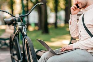A young man seated on a bench in a park engages in a phone call while using a laptop. His bike rests nearby, showcasing a blend of technology and lifestyle in a relaxing outdoor setting. photo