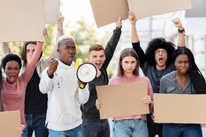 Furious multiracial students with empty placards having strike or demonstration on the street. Motivated african american man with loudspeaker chanting slogans over demonstrating crowd outdoors photo