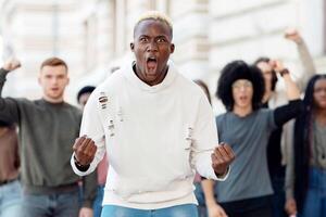 Multiracial group of activists chanting slogans, raising fists up, making strike on the street. Furious black guy guiding millennial men and wome, protesting against discrimination photo