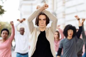 Young man leading multiracial group of activists, crossing fists up, showing unity and strength, millennial strikers different nationalities walking by street, making riot against quarantine rules photo