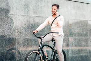Urgent call, work on move and active, sports lifestyle. Smiling attractive millennial guy manager speaks on phone and rides on bike at city, on gray street wall background, outdoor, free space photo