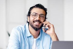Cheerful Indian Freelancer Guy Drinking Coffee And Talking On Cellphone While Having Break During Work With Laptop Computer At Home Office, Speaking With Friends, Resting At Workplace, Free Space photo
