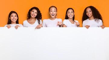 Excited Multicultural Ladies Holding Empty Paper Board Advertising Your Text Posing Standing Over Yellow Background In Studio, Smiling To Camera. Mockup, Panorama photo