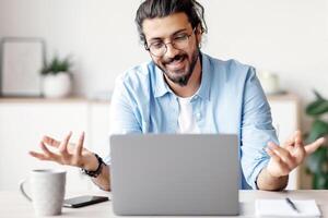 Online Tutoring. Friendly Indian Man Having Conference With Laptop, Teaching Foreign Languages, Talking And Gesturing At Camera During Web Call, Sitting At Desk In Home Office, Copy Space photo