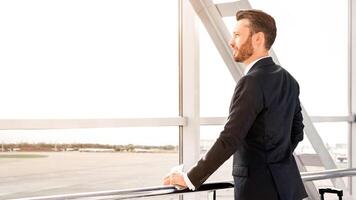 Side view of successful businessman with suitcase standing next to window at airport, looking at airplane, copy space. Wealthy man in black suit waiting for departure, looking through the window photo
