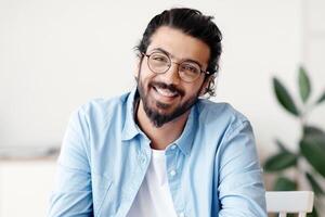 Portrait Of Happy Western Guy In Eyeglasses Sitting At Desk In Home Office, Smiling At Camera, Enjoying Remote Work, Cheerful Indian Freelancer Man Posing While Working With Laptop Indoors, Free Space photo
