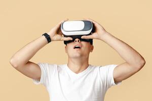 Amazing virtual world and modern technology for study and entertainment. Surprised young man with open mouth in virtual reality glasses looking up at empty space, isolated on beige background photo