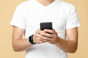 Social networks, blogs and mobile app. Smiling handsome young blogger student in white t-shirt with smart watch in glasses hold phone and surfing in internet isolated on light background, cropped photo