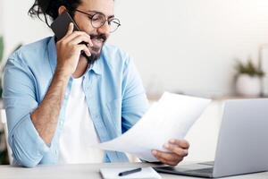 Handsome Indian Freelancer Guy Talking On Cellphone And Working With Papers At Home Office, Discussing Project Budget With Client, Checking Documents And Financial Reports, Sitting At Desk, Copy Space photo