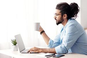 Young Eastern Freelancer Guy Working On Laptop At Home And Drinking Coffee, Sitting At Desk Using Computer And Having Hot Drink, Millennial Arab Man Enjoying Remote Work And Distance Job, Side View photo