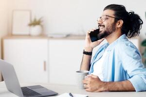 Cheerful Indian Freelancer Guy Resting With Coffee And Cellphone At Desk In Home Office, Talking On Mobile Phone With Friends And Smiling, Having Break At Work, Enjoying Working Remotely, Copy Space photo