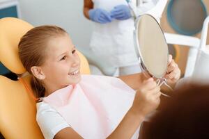 Treat bad teeth and snow-white smile after procedure. Happy little child in dental chair looks in mirror and evaluates work of medical team in white coats, protective masks and rubber gloves photo