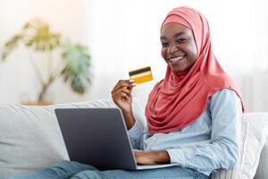 Online Shopping. Happy black muslim woman in hijab using laptop and credit card at home, purchasing in internet, paying for utilities, enjoying easy e-banking payments, transferring money, copy space photo