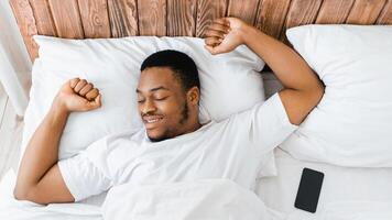 Good Morning Concept. Wellslept Black Guy Awakening In Bed Stretching Hands After Sleeping In Bedroom At Home. Man Waking Up In Bed, Recreation And Rest. Panorama, High-Angle View photo