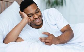 Cheerful Black Man Using Phone Browsing Internet And Networking Texting On Cellphone Lying In Bed At Home. New Mobile Application For Smartphone Advertisement Concept. Selective Focus photo