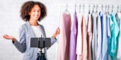 Modern technology for work online from home during self isolating. Blurred happy african american business woman presenting collection of clothes and recording on smartphone, close up photo