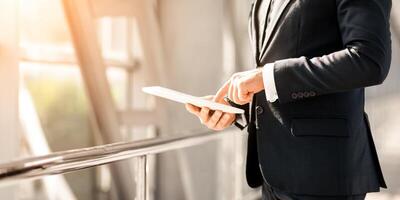 Unrecognizable entrepreneur with digital tablet standing next to window in airport, using nice mobile application for business while waiting for flight, panorama with copy space, sun flare, cropped photo