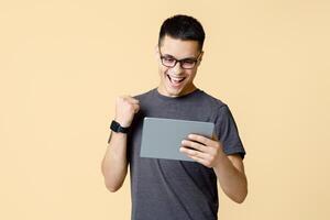 Online victory, excellent mark or review and good result. Happy young guy student in glasses with open mouth looks in tablet makes win gesture, isolated on beige background, studio shot, free space photo