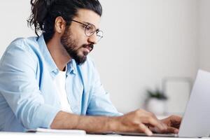 Young Eastern Employee Working On Laptop At Desk In Office, Busy Arab Male Entrepreneur Sitting At Workplace, Typing On Keyboard And Looking At Computer Screen, Checking Online Project, Free Space photo