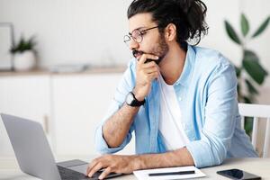 Thoughtful Indian Freelancer Working With Laptop At Home Office, Sitting At Desk Looking At Computer Screen, Thinking About Project Solution, Browsing Information In Internet, Copy Space photo