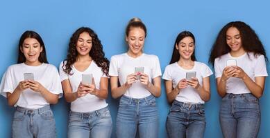 Five Happy Multiracial Females Using Phones Texting Networking In Social Media Standing On Blue Background. Studio Shot Of Millennial Women Using Mobile Application Posing Together. Panorama photo
