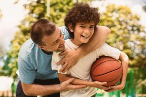 Enjoying Game. Portrait of cheerful father and son playing basketball together, smiling positive boy holding ball and looking at camera, excited man embracing him from behind and trying to take ball photo