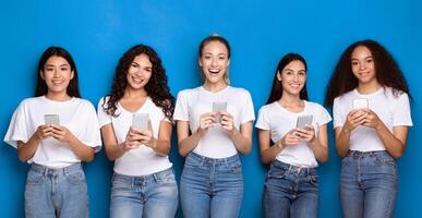 Five Multiracial Women Using Smartphones Posing Standing Over Blue Studio Background, Smiling To Camera. Mobile Phone Users Texting In Social Media And Using Applications. Panorama photo