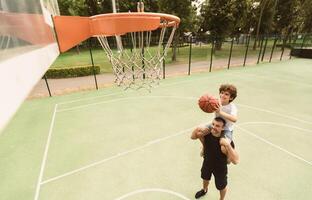 Support. High angle view of man holding his curly son on shoulders, helping him to score a basket on playground photo