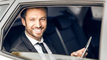Smiling handsome businessman in suit sitting in car, looking through window, using digital tablet photo