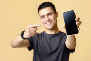 Great review, ad and advice from blogger. Happy confident guy student in brown t-shirt recommends app and points finger at phone with blank screen, isolated on beige background, studio shot photo