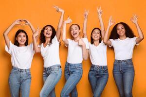 Group Of Multiracial Ladies Gesturing Different Positive Signs And Symbols Raising Hands Posing Over Yellow Studio Background, Smiling To Camera. Females Showing Okay, V-Sign And Thumbs Up Gestures photo