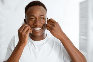 Happy African American Man Flossing Teeth Standing In Modern Bathroom At Home, Smiling Looking At Camera. Teeth Cleaning With Floss, Oral Toothcare Hygiene Routine Concept photo