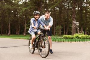 Leisure Concept. Portrait of cheerful adult father teaching his son how to ride a bicycle at public park, happy loving family spending time together on weekend, having fun outdoors in the summer photo