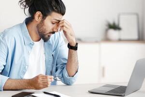 Eyes Fatigue. Stressed Indian Freelancer Man Tired After Working On Laptop Computer In Home Office, Massaging Nosebridge,Taking Off Glasses, Having Headache Or Problems At Work, Free Space photo