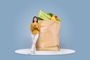 A happy customer stands next to a large paper bag overflowing with fresh vegetables and fruits while using a smartphone. The setting reflects modern shopping habits amid current lifestyles. photo