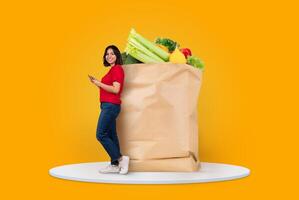 A customer stands beside a large paper bag overflowing with fresh vegetables and fruits, while checking their smartphone. This represents modern shopping habits and remote delivery options. photo