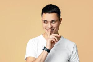 Playful young guy in white t-shirt keeps finger on lips, looking away at copy space and smiling, beige studio background photo