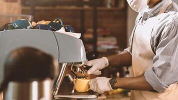 New normal and customer service during covid-19 lockdown. Millennial african american man in apron, protective mask and gloves makes latte in yellow cup on coffee machine at cafe interior, cropped photo
