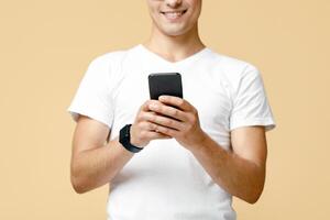 Surfing on smartphone or write in online blog. Attractive smart smiling young man in white t-shirt typing at phone and reading message, isolated on beige background, studio shot, free space photo