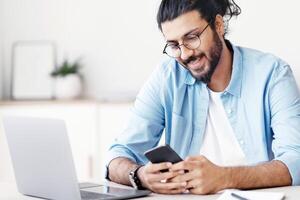 Handsome Eastern freelancer guy sitting at desk at home office and using smartphone, messaging with friends, checking social media or browsing new app, enjoying mobile communication, free space photo