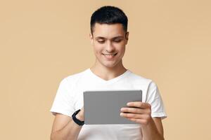 Device for work, entertainment, rest and communication online. Happy guy uses tablet for typing to friends, plays in game and chatting isolated on beige background, studio shot, free space photo