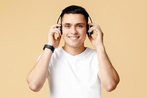 Relax at home or walking and playlist in online app. Cheerful guy student in white t-shirt and headphones listens to favorite music, isolated on beige background, studio shot, free space photo