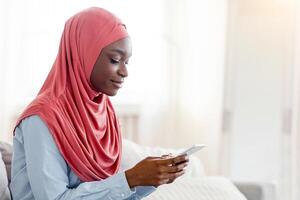 Happy black muslim woman surfing internet on smartphone while resting on couch at home, relaxing in cozy living room with cellphone, sitting on sofa and browsing social networks, side view, copy space photo